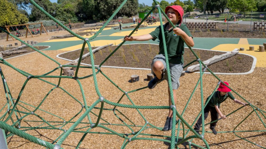 Two children climb a green rope climbing frame at a playground on a sunny day.