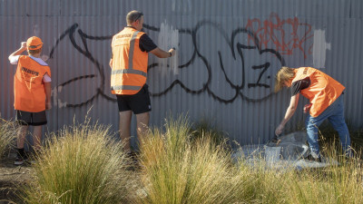 three people paint over some graffiti on a fence